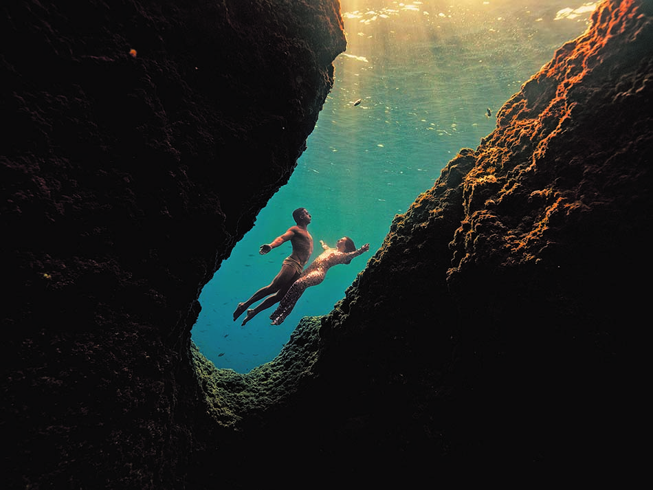 Zwei Taucher erkunden einen Unterwasserpassage, umgeben von Licht und Meeresfelsen.